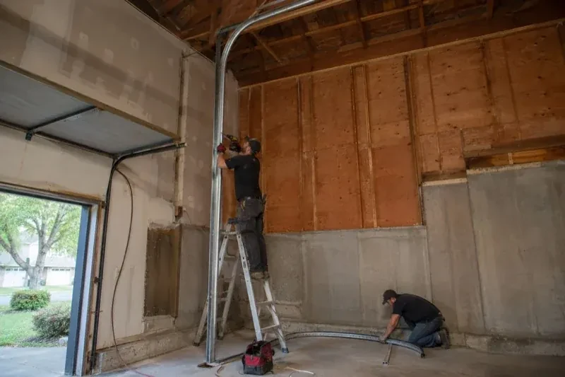 One person on a stepladder working beside a tall curved garage-door track while another kneels beside loose track sections in an unfinished garage. — High-Lift Garage Door Conversion, Vaughan